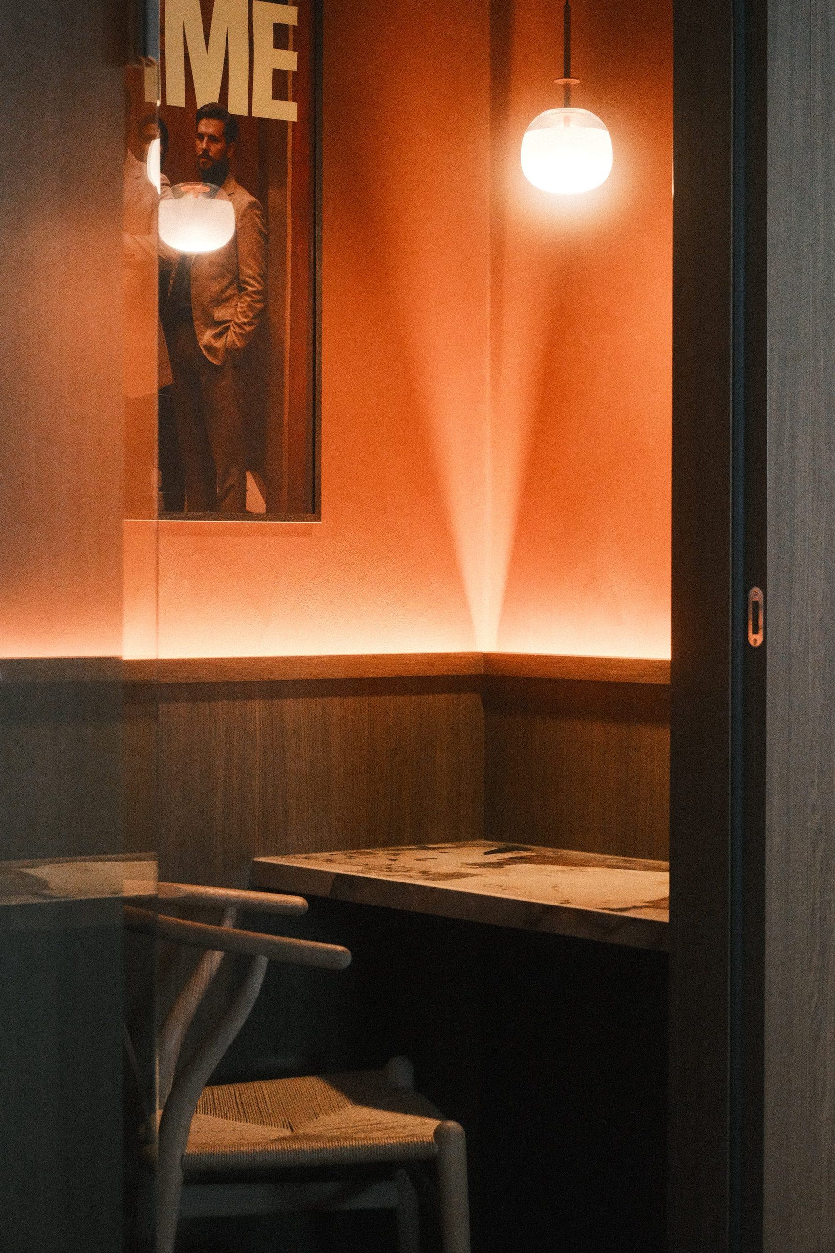 Phone booth with a small marble table and a chair, a glass globe pendant and a warm line of light against an orange wall