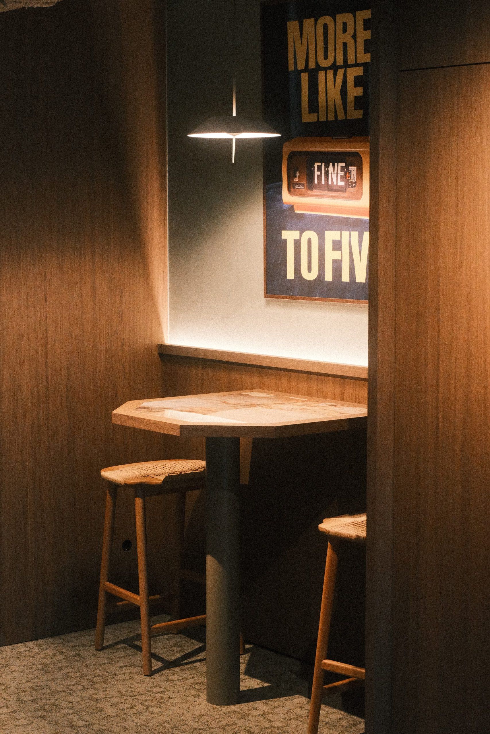 Niche with a high table and stools beneath a flat pendant, with a line of light above the timber panelling and a poster on the wall