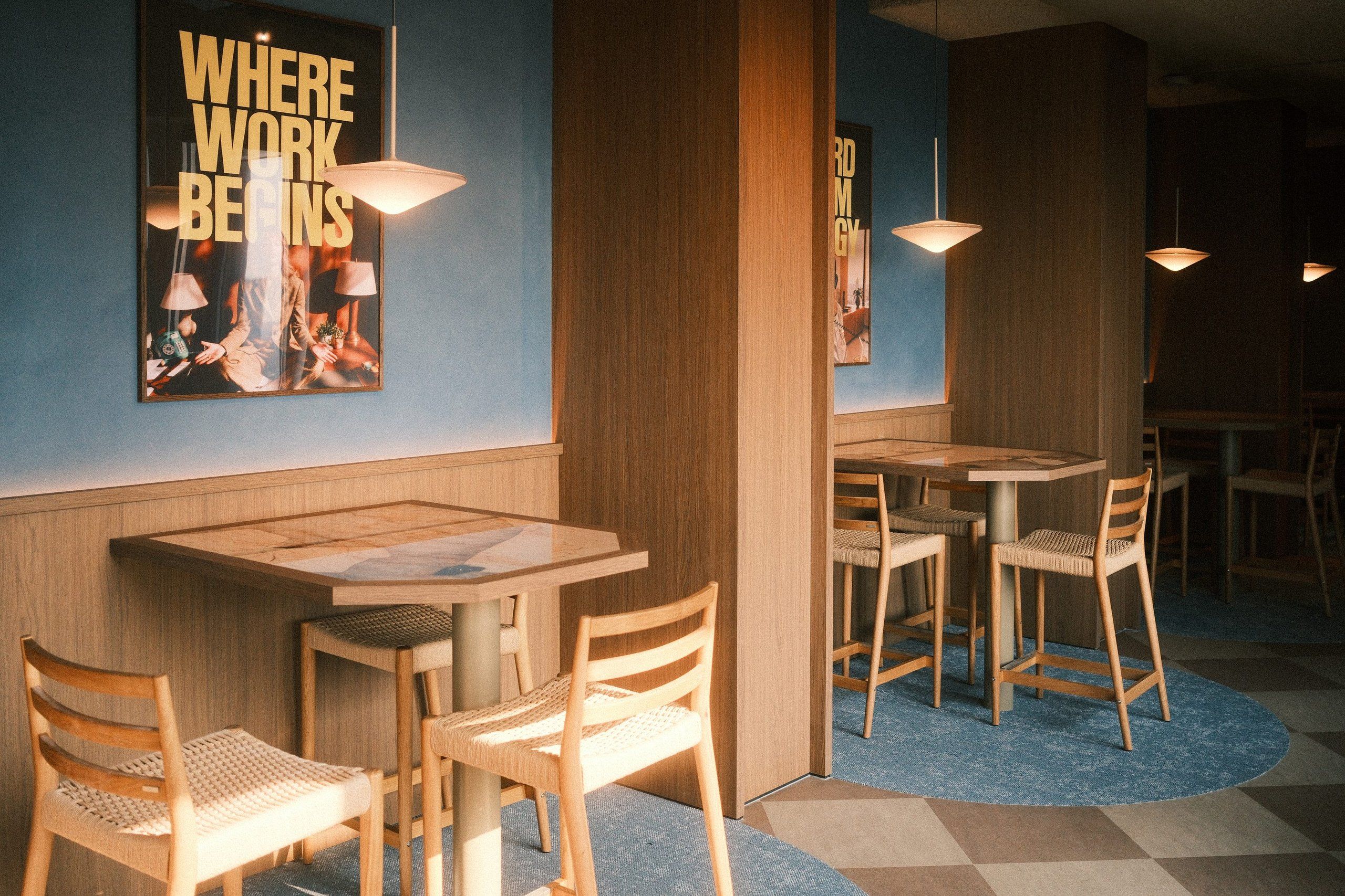 Timber high tables and stools in afternoon light beneath flat pendants, against a blue wall with a poster