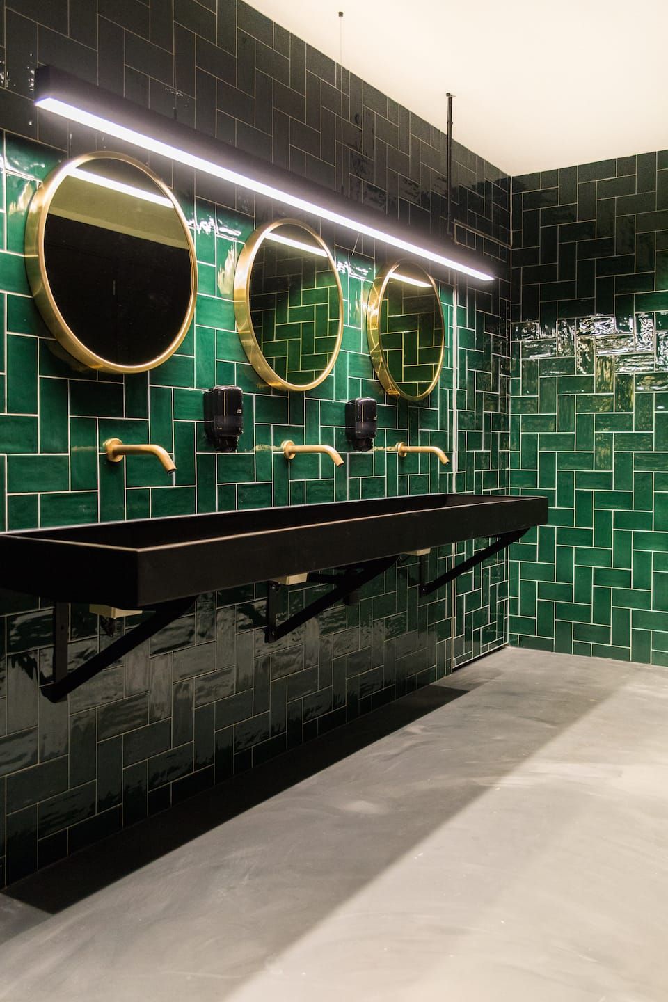 Green glazed tile restroom with round brass mirrors and a black trough sink
