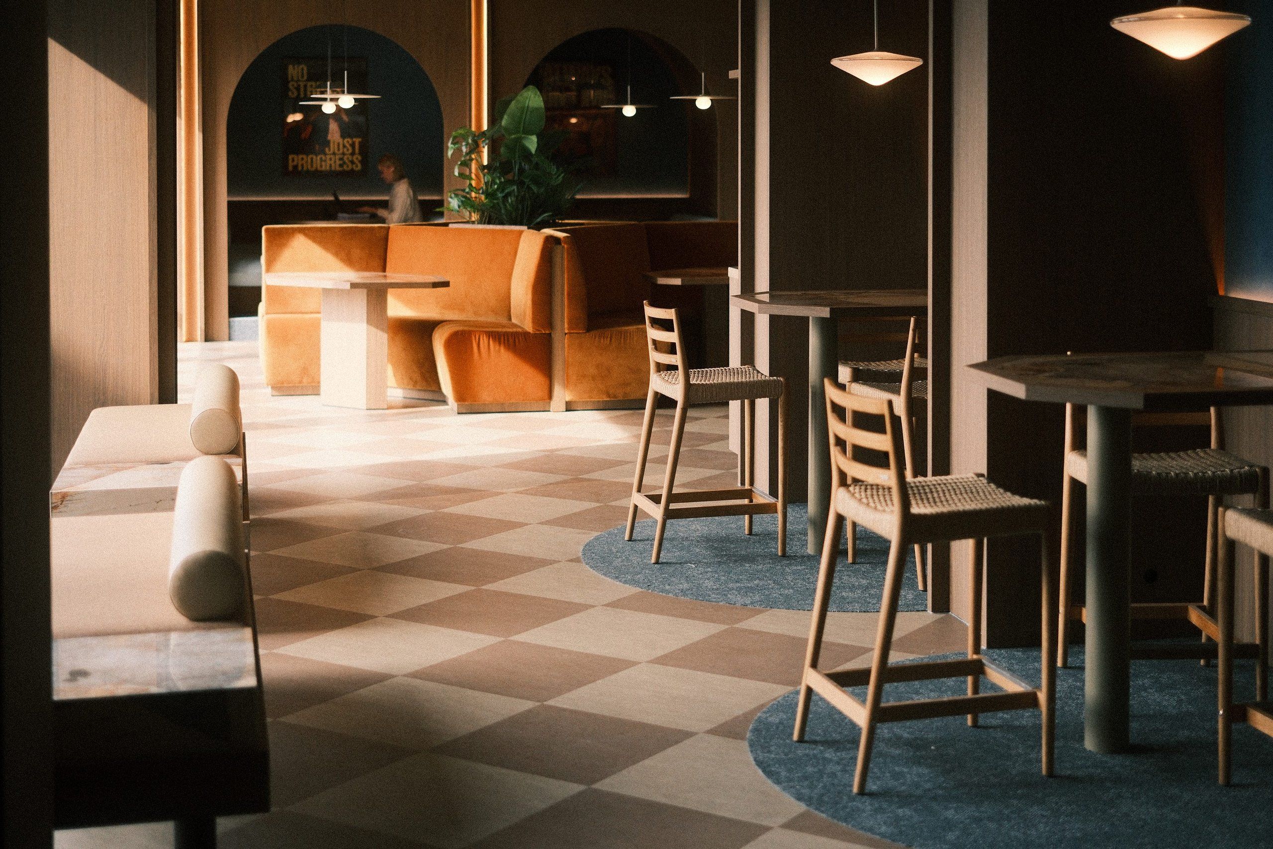 Work spots at timber high tables on a checkered floor beneath flat pendants, with an orange velvet banquette in an arch at the back, in the Officina office